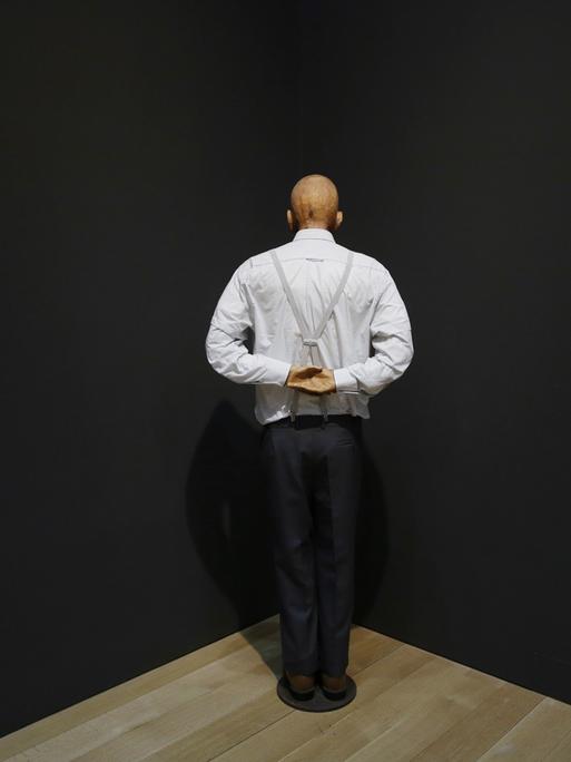 Die Installation "Martin, ab in die Ecke und schäm dich". Zu sehen ist eine Figur von hinten, die in einer schwarzen Raumecke steht und die Hände auf dem Rücken zusammenlegt. Die Figur trägt ein weißes Hemd und graue Hosenträger. 