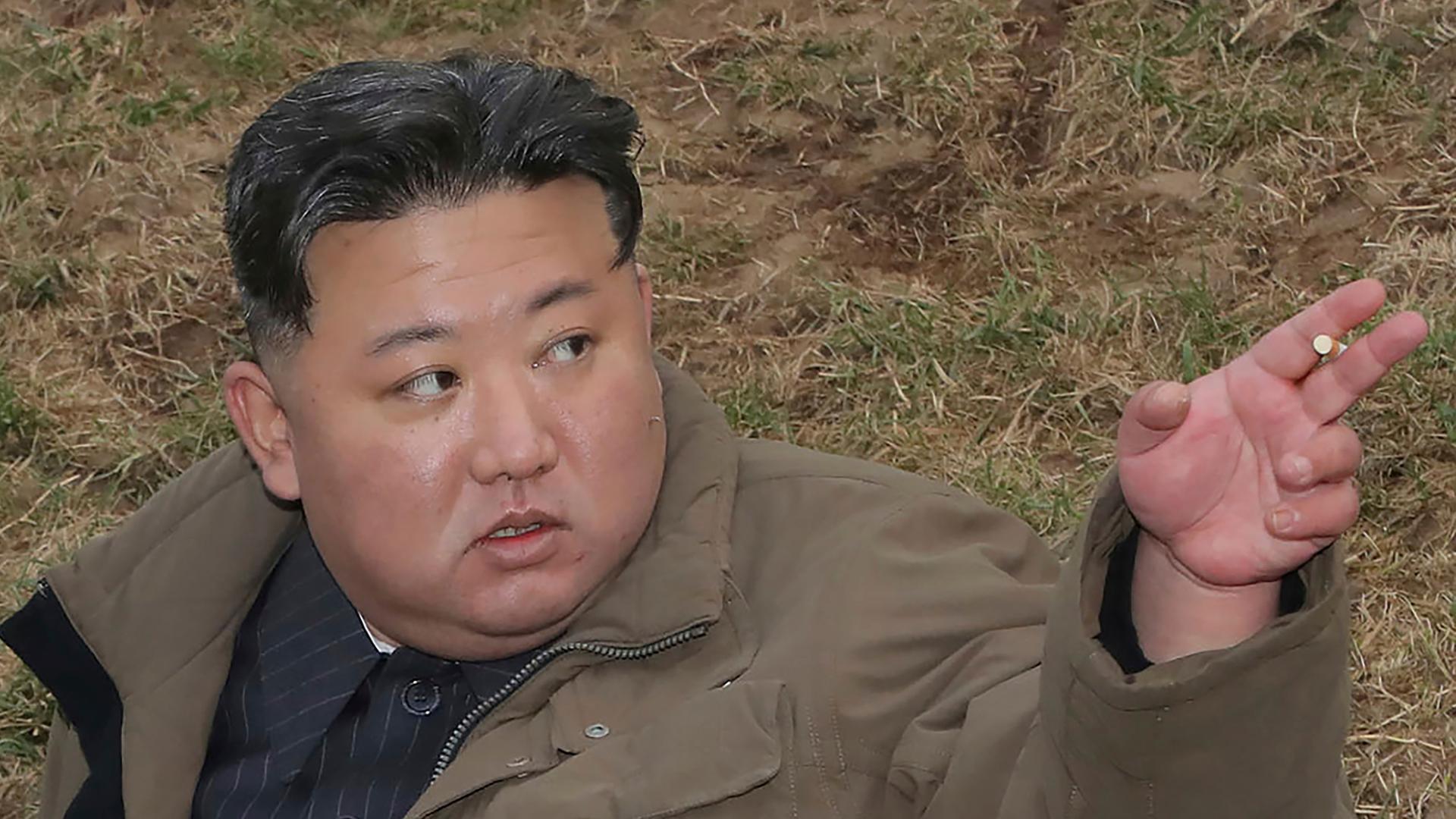 Kim in einer brauene Jacke steht auf einem braunen Rasen und deutet mit der linken Hand nach oben. Er ist von schräg oben fotografiert.