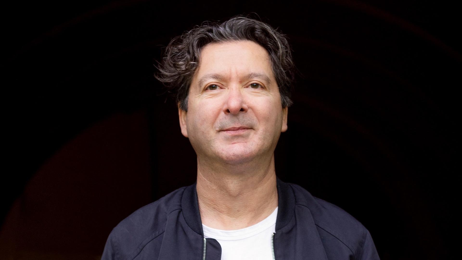 Mustafa Akça vor schwarzem Hintergrund in blauer Jacke.