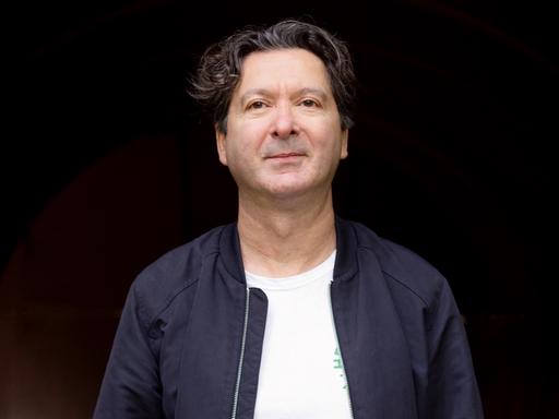 Mustafa Akça vor schwarzem Hintergrund in blauer Jacke.