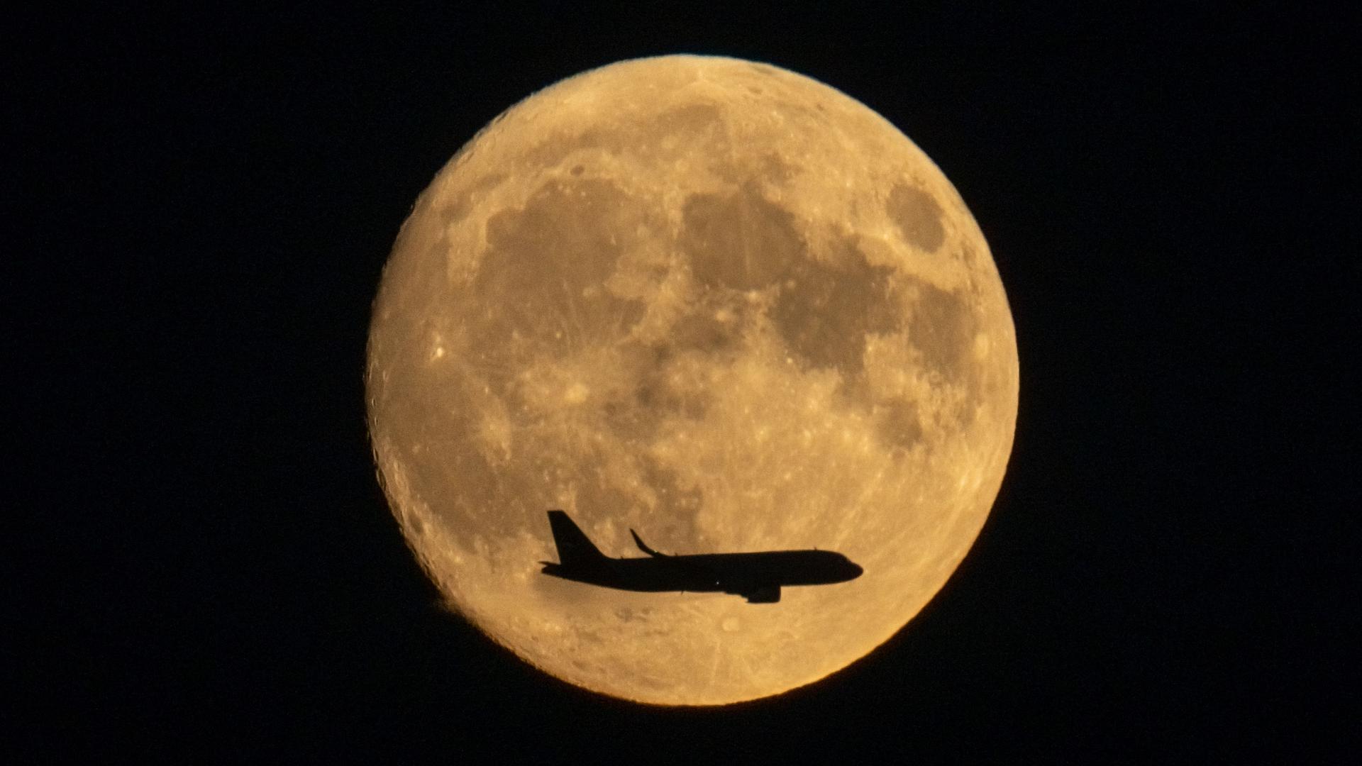 Ein Flugzeug im Landeanflug auf den Frankfurter Flughafen fliegt am Mond vorbei, der gelblich schimmernd am späten Abendhimmel zu sehen ist.