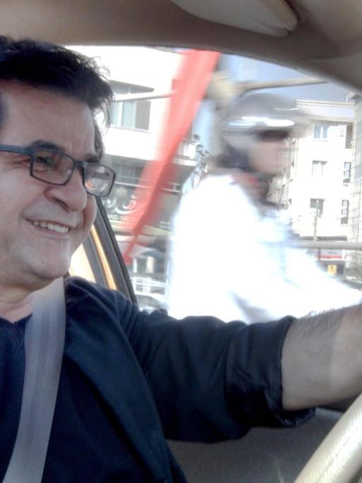 Jafar Panahi sitzt in einem Auto am Steuer und lächelt. 