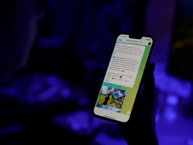 Der Telegram-Messengerdienst ist auf dem Display eines Smartphones zu sehen, dass eine Person in der Hand hält. Die Person ist nur als Schattenumriss zu erkennen, der Hintergrund schimmert bläulich.