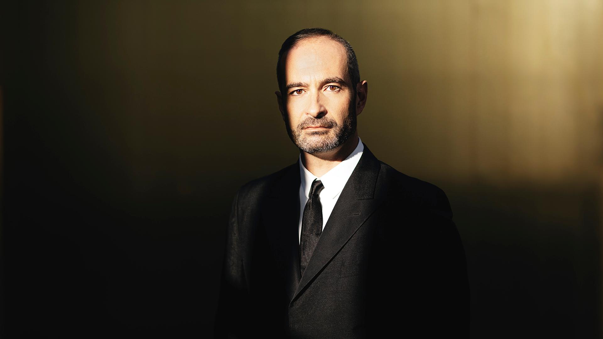 Ein Mann im schwarzen Anzug, kurzen schwarzgrauen Haaren und einem drei Tage Bart blickt in die Kamera. 