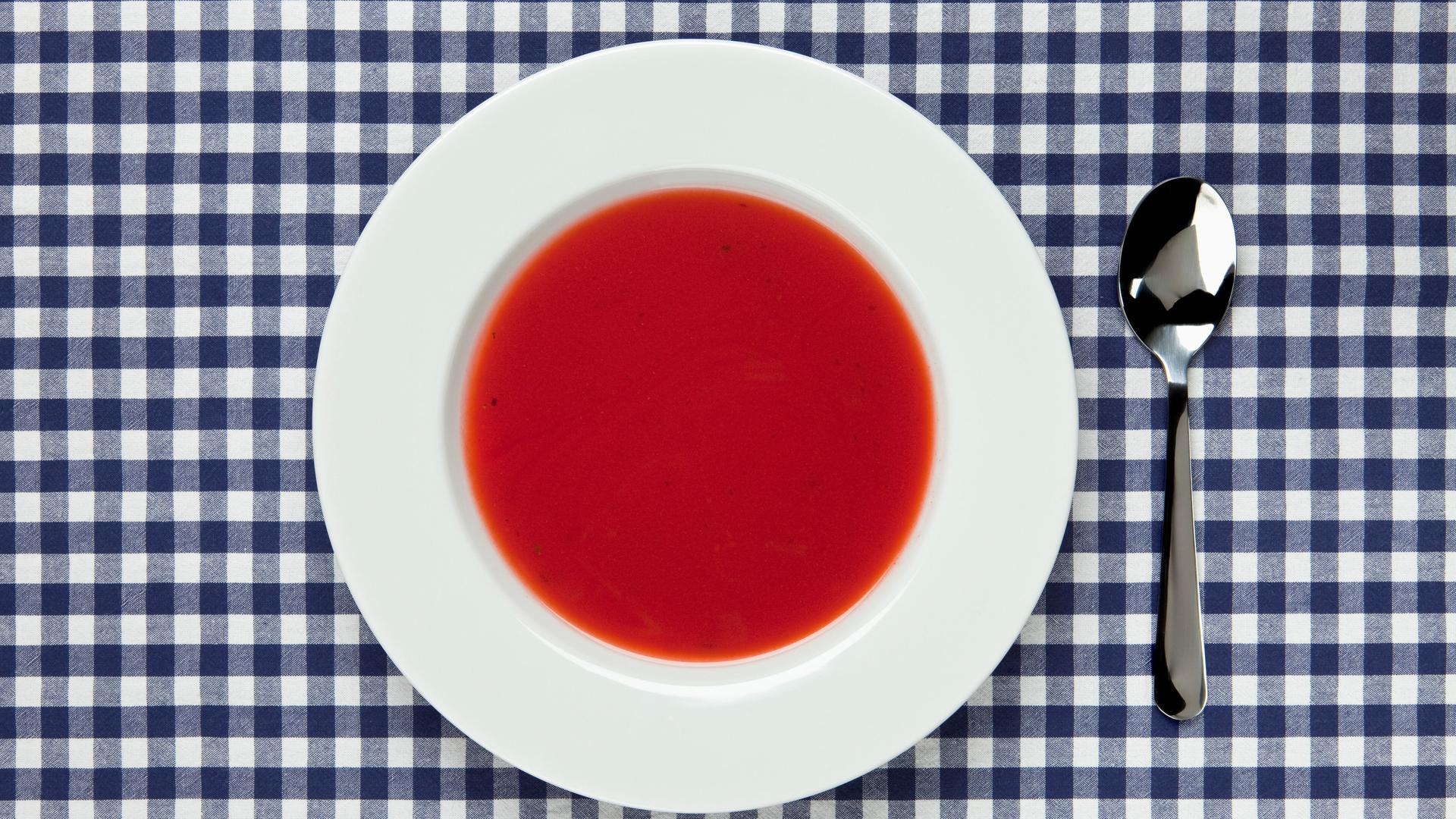 Auf einem Tisch mit blau-karierter Tischdecke steht ein weisser Teller mit Tomatensuppe. Daneben ein Löffel.