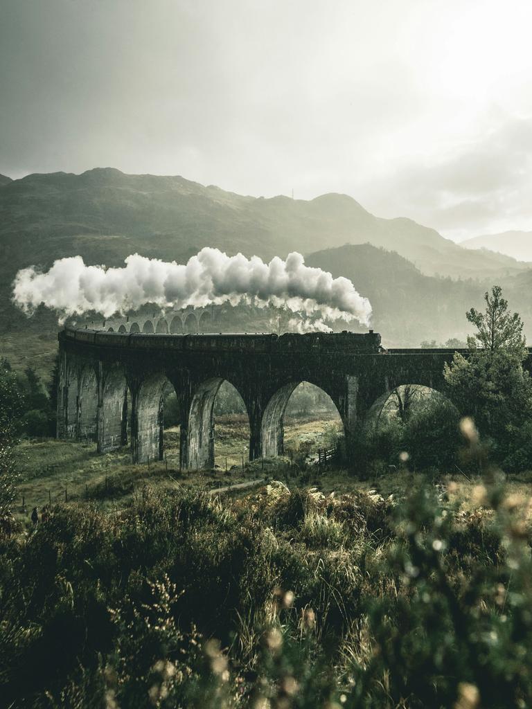 Eine alte Dampflok fährt über ein Viadukt durch eine hügelige Naturlandschaft.