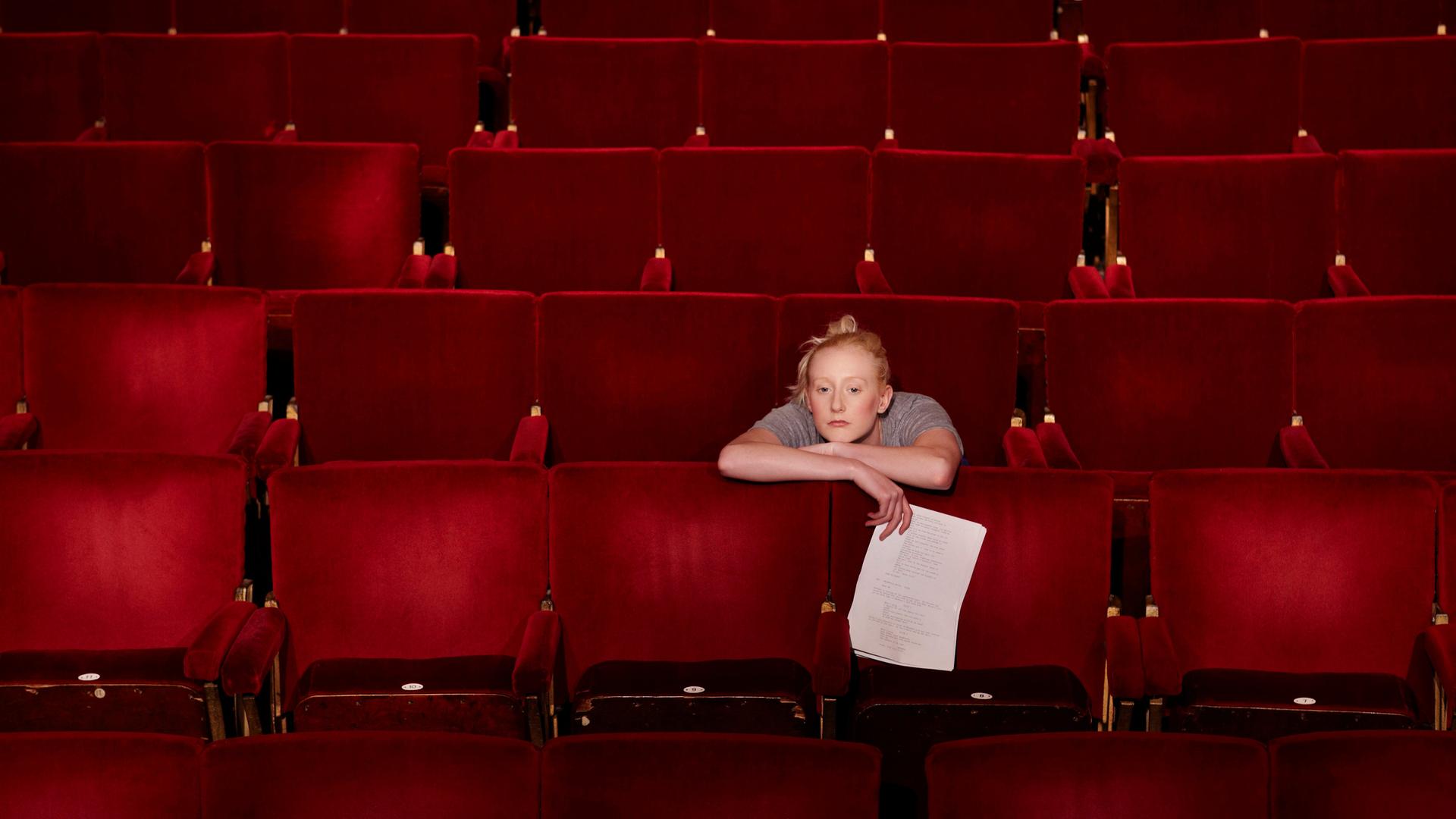 Eine blonde Frau mit lockerem Dutt sitzt in einem Theatersaal über die Lehne des roten Klappsitzes vor ihr gelehnt und hält ein Blatt in der rechten Hand. Eine blonde Frau mit lockerem Dutt sitzt in einem Theatersaal über die Lehne des roten Klappsitzes vor ihr gelehnt und hält ein Blatt in der rechten Hand.