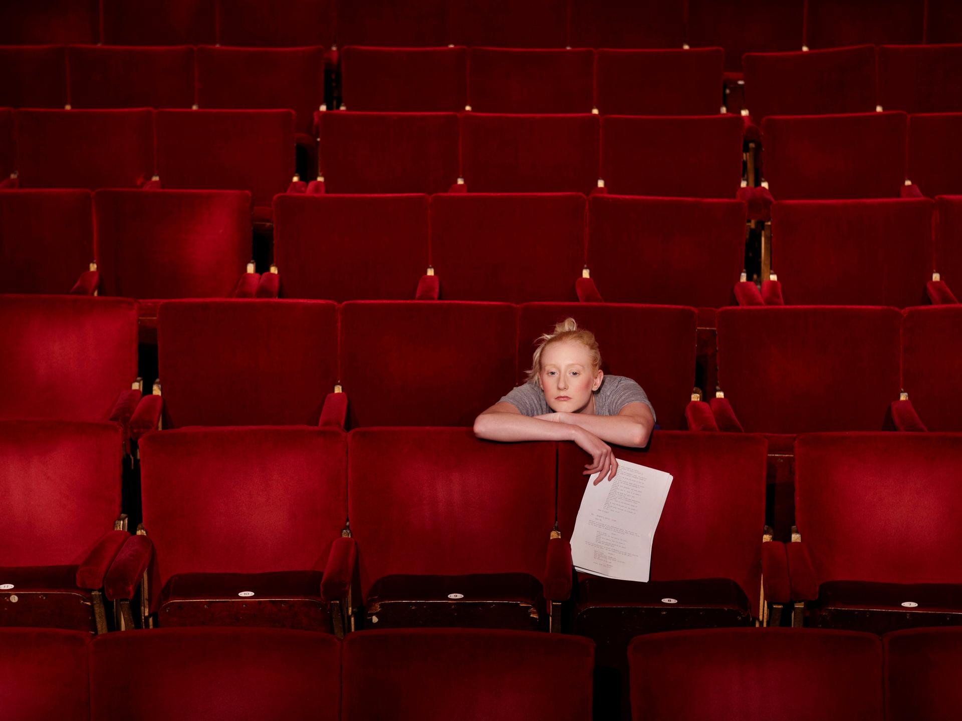 Eine blonde Frau mit lockerem Dutt sitzt in einem Theatersaal über die Lehne des roten Klappsitzes vor ihr gelehnt und hält ein Blatt in der rechten Hand.