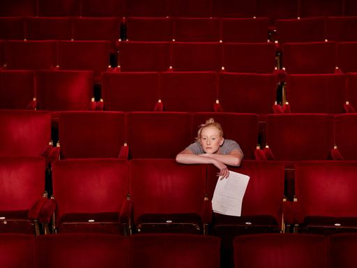 Eine blonde Frau mit lockerem Dutt sitzt in einem Theatersaal über die Lehne des roten Klappsitzes vor ihr gelehnt und hält ein Blatt in der rechten Hand.