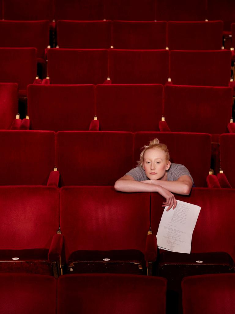 Eine blonde Frau mit lockerem Dutt sitzt in einem Theatersaal über die Lehne des roten Klappsitzes vor ihr gelehnt und hält ein Blatt in der rechten Hand.