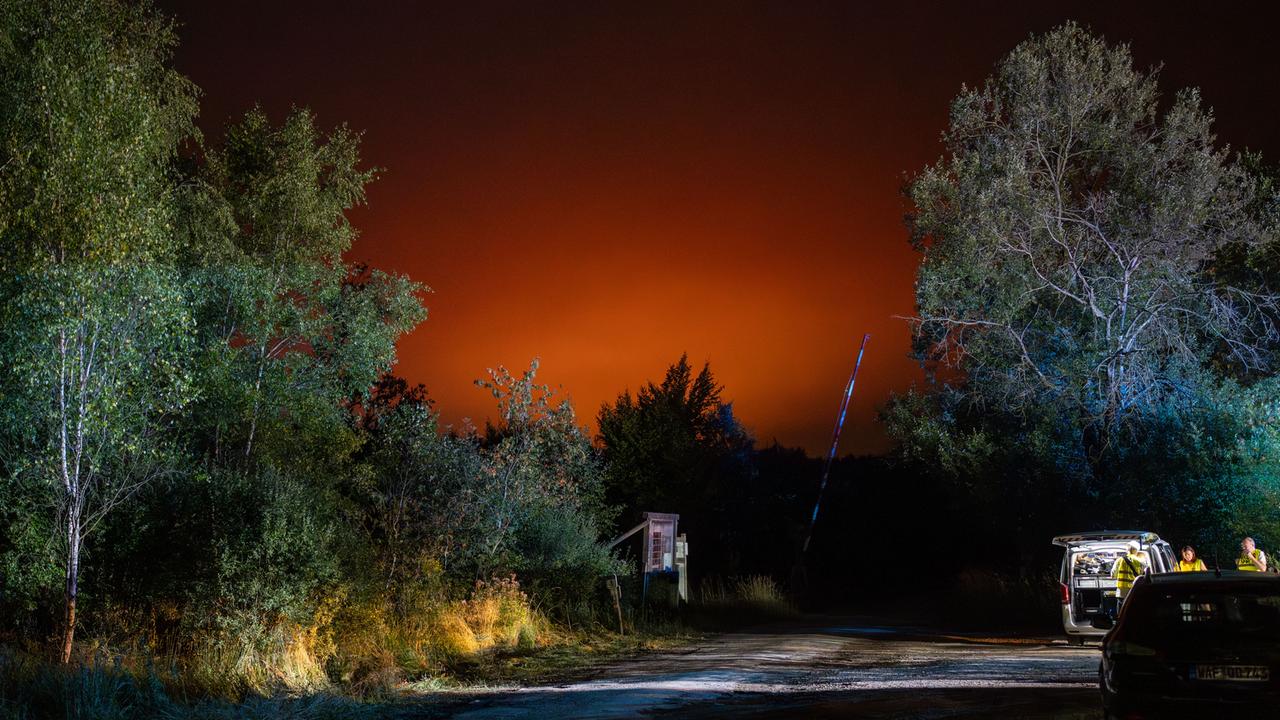 Rettungskräfte stehen auf einem Waldparkplatz. Der Nachthimmel ist rot erhellt vom weiter entfernten Waldbrand. 