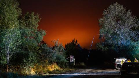 Rettungskräfte stehen auf einem Waldparkplatz. Der Nachthimmel ist rot erhellt vom weiter entfernten Waldbrand. 