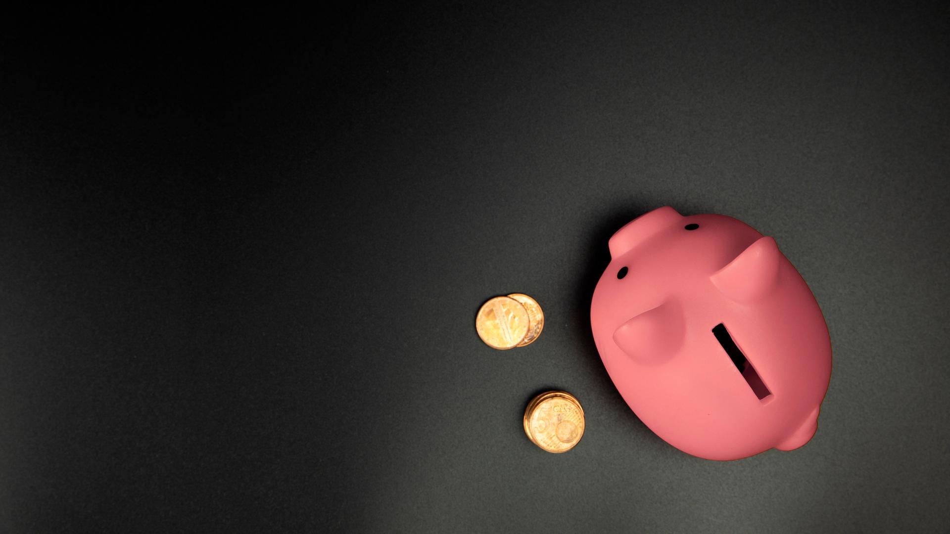 Ein pinkes Sparschwein steht auf einem schwarzen Untergrund, rechts davon liegen Cent-Münzen.