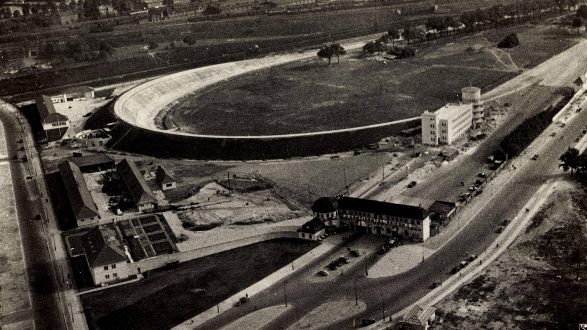 Schwarzweiß-Foto von 1935 mit Blick von oben auf die Nordschleife der Avus Berlin. Man sieht die Steilkurve und rechts ein Gebäude, das als Tribüne und Hotel bei Autorennen diente. 