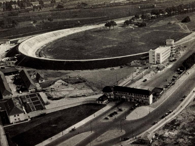 Schwarzweiß-Foto von 1935 mit Blick von oben auf die Nordschleife der Avus Berlin. Man sieht die Steilkurve und rechts ein Gebäude, das als Tribüne und Hotel bei Autorennen diente. 