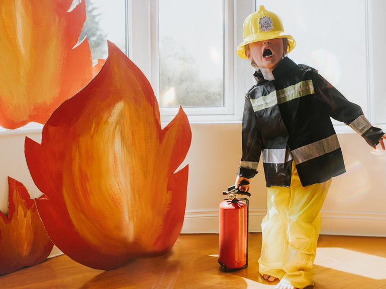 Ein Kind verkleidet als Feuerwehr, mit Helm, Anzug, Feuerlöscher und Flammen aus Pappe.