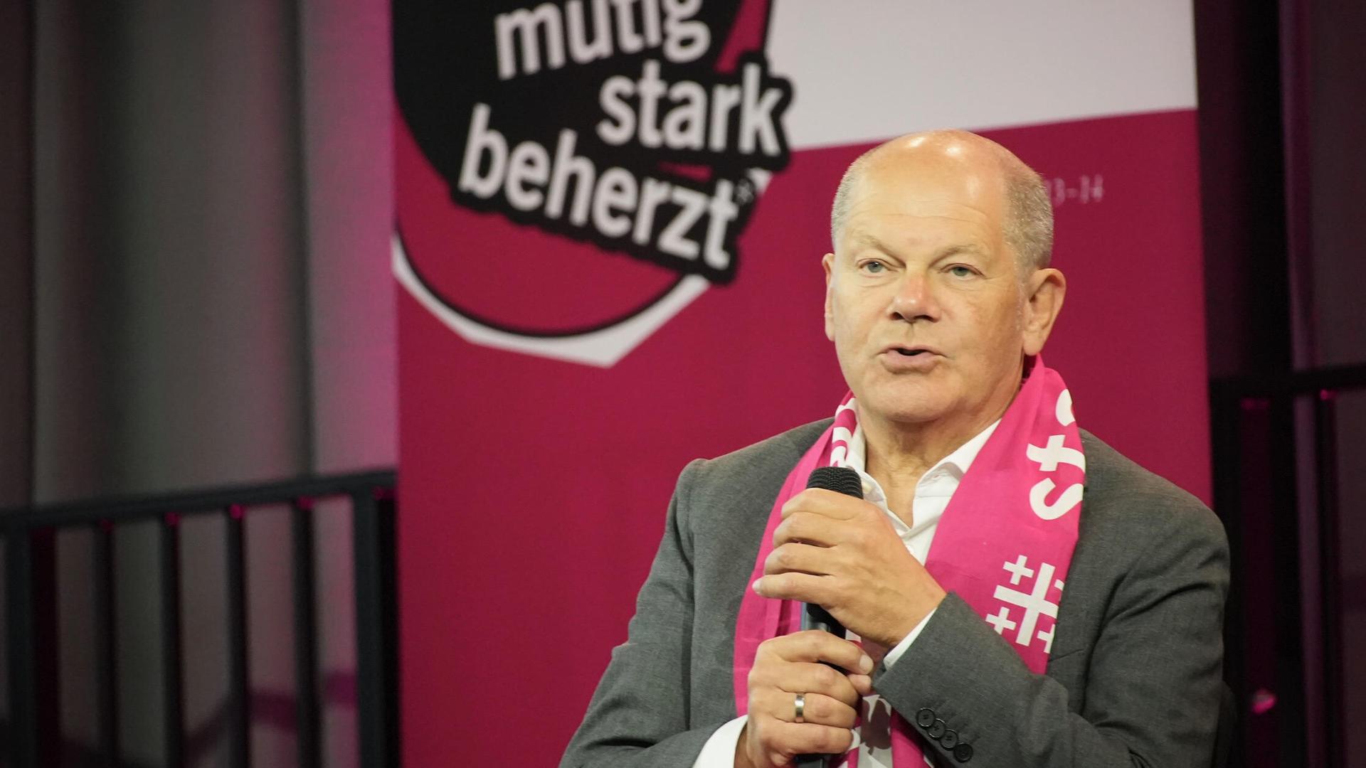 Scholz auf einer Bühne, hält das Mikrofon vor sich. Hinter ihm als Transparent das Motto des Kirchentages: "mutig, stark, beherzt"