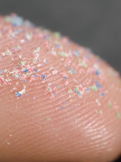 Auf einer Fingerspitze sind Plastikpartikel im bunten Farben zu sehen.
