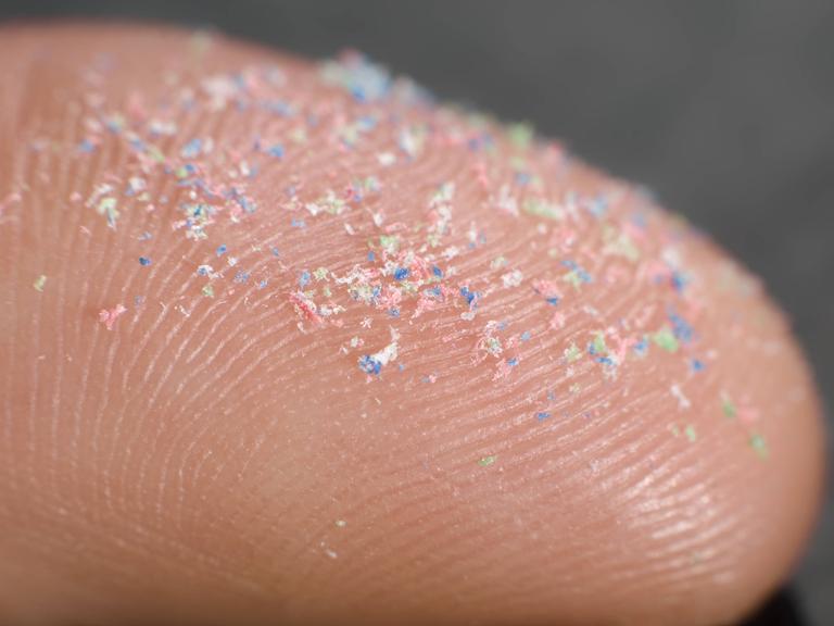 Auf einer Fingerspitze sind Plastikpartikel im bunten Farben zu sehen.