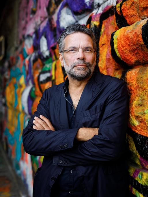 Robert Pfaller steht in dunklem Sakko und verschränkten Armen vor einer farbig besprühten Wand und schaut freundlich in die Kamera.