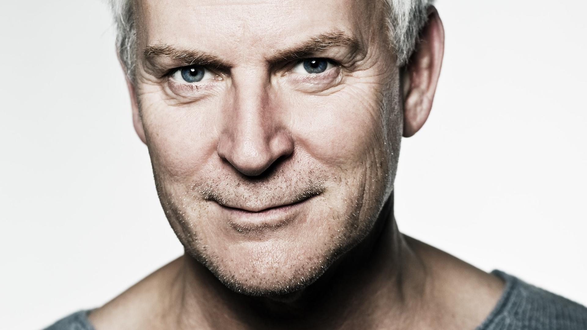 Ein Mann mit grauem Haar und Dreitagebart schaut schmunzelnd in die Kamera. Er trägt einen dunkelgrauen Pullover, der dieselbe Farbe wie seine Augen hat. Der Hintergrund ist weiß. Ein Mann mit grauem Haar und Dreitagebart schaut schmunzelnd in die Kamera. Er trägt einen dunkelgrauen Pullover, der dieselbe Farbe wie seine Augen hat. Der Hintergrund ist weiß.