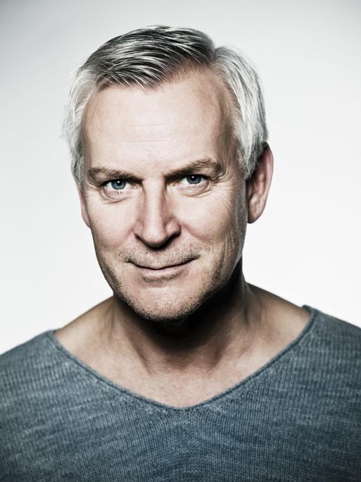 Ein Mann mit grauem Haar und Dreitagebart schaut schmunzelnd in die Kamera. Er trägt einen dunkelgrauen Pullover, der dieselbe Farbe wie seine Augen hat. Der Hintergrund ist weiß.