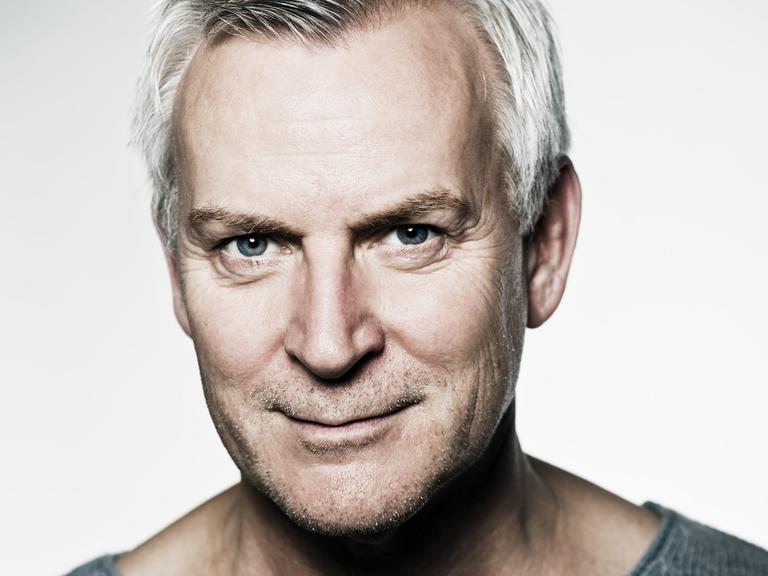 Ein Mann mit grauem Haar und Dreitagebart schaut schmunzelnd in die Kamera. Er trägt einen dunkelgrauen Pullover, der dieselbe Farbe wie seine Augen hat. Der Hintergrund ist weiß.