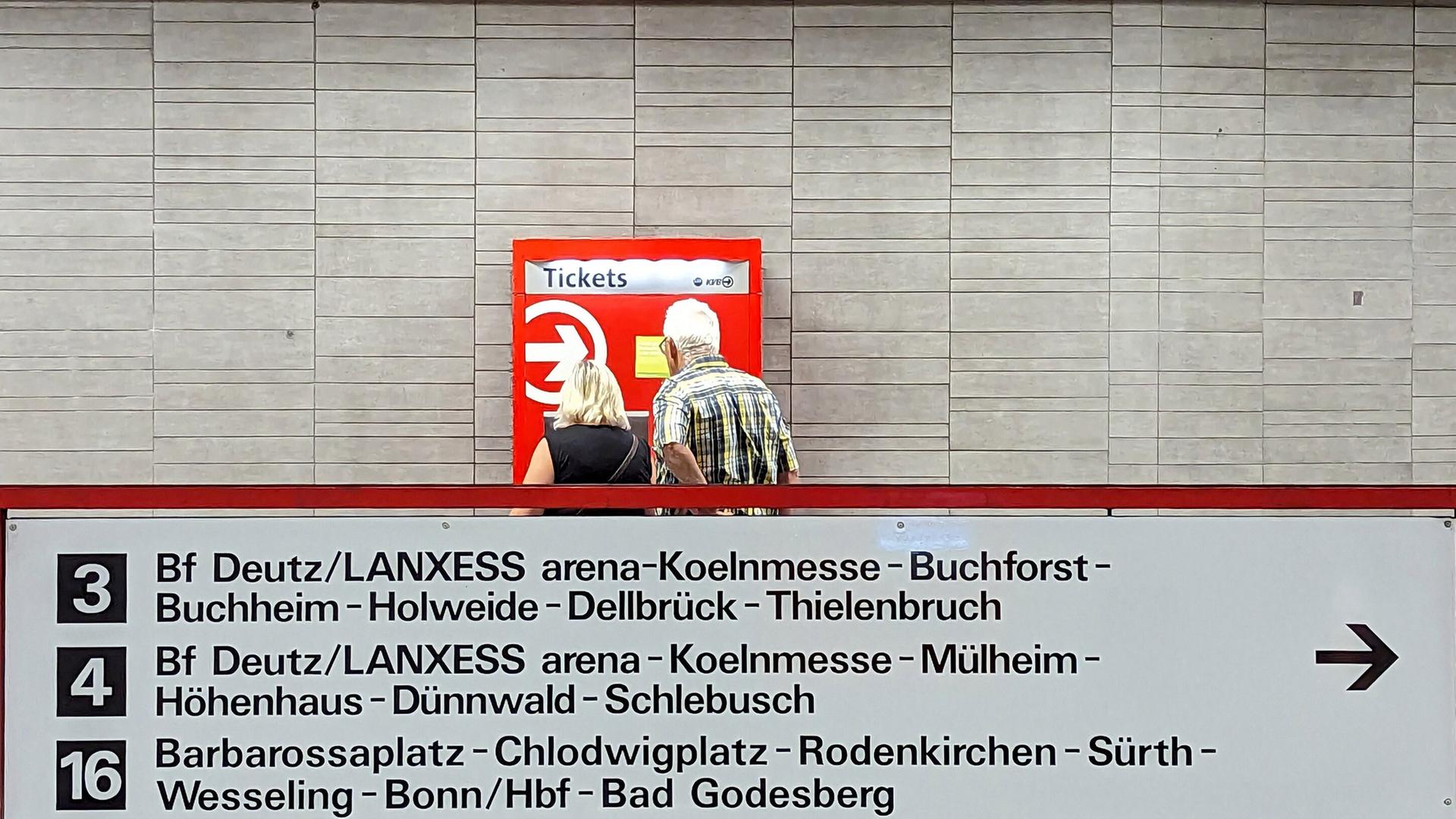 Eine Frau und ein Mann stehen in Köln vor einem rot-weißen Fahrkartenautomaten. Vor ihnen ist eine Tafel mit verschiedenen ÖPNV-Verbindungen.