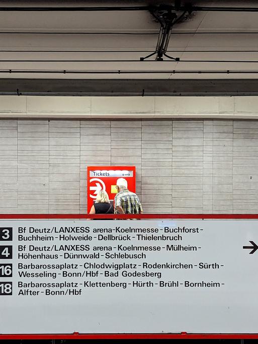 Eine Frau und ein Mann stehen in Köln vor einem rot-weißen Fahrkartenautomaten. Vor ihnen ist eine Tafel mit verschiedenen ÖPNV-Verbindungen.