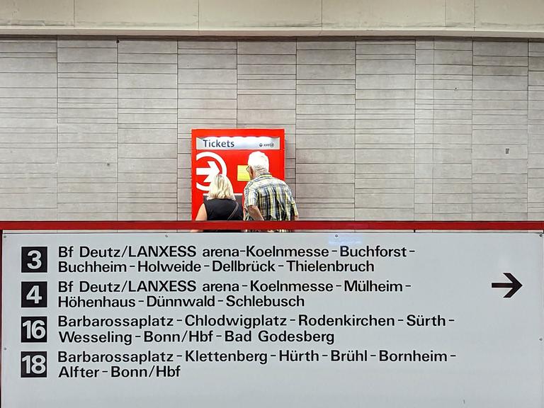 Eine Frau und ein Mann stehen in Köln vor einem rot-weißen Fahrkartenautomaten. Vor ihnen ist eine Tafel mit verschiedenen ÖPNV-Verbindungen.