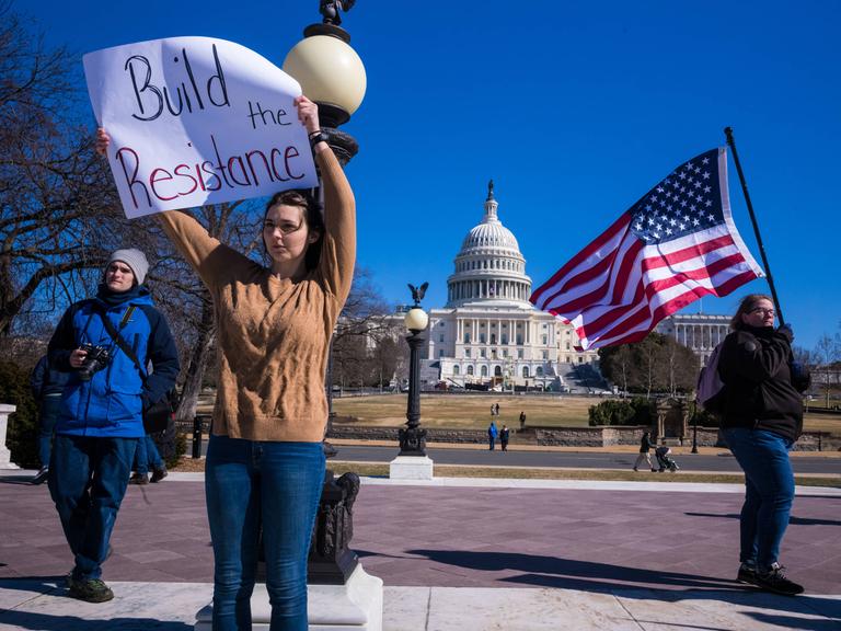 Drei Menschen, mit Flagge und einem bemalten Schild protestieren gegen Trump vor dem Kapitolgebäude in Washington