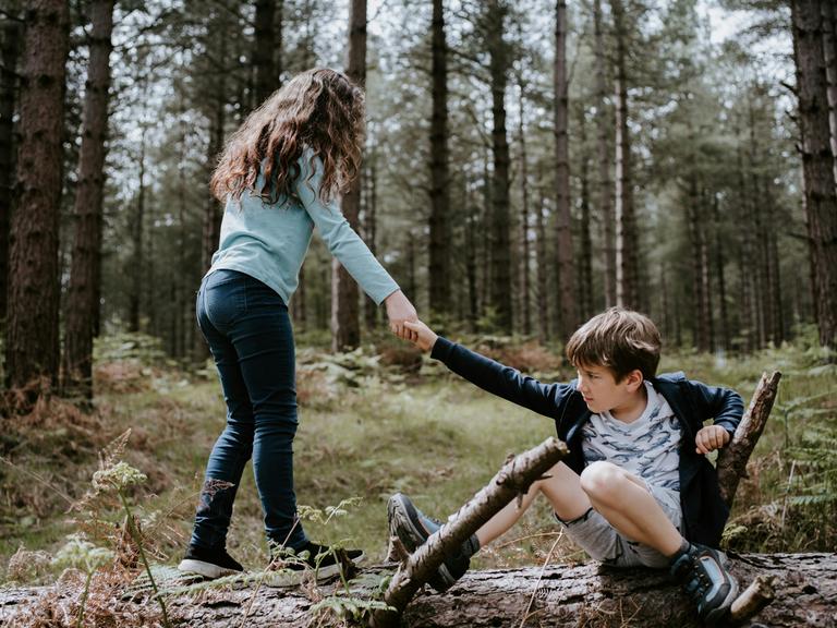 Zwei Kinder im Wald. Der Junge hockt am Boden. Das Mädchen hilft ihm auf.