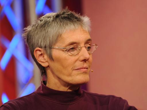 Petra Morsbach sitzt in einem Fernsehstudio und schaut zur Seite. Sie hat kurze Haare und trägt eine Brille.