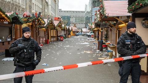 Auf dem Weihnachtsmarkt stehen Polizisten hinter einer Absperrung mit Flatterband. Im Hintergrund stehen die Buden des Weihnachtsmarkts, Müll liegt auf der Straße.