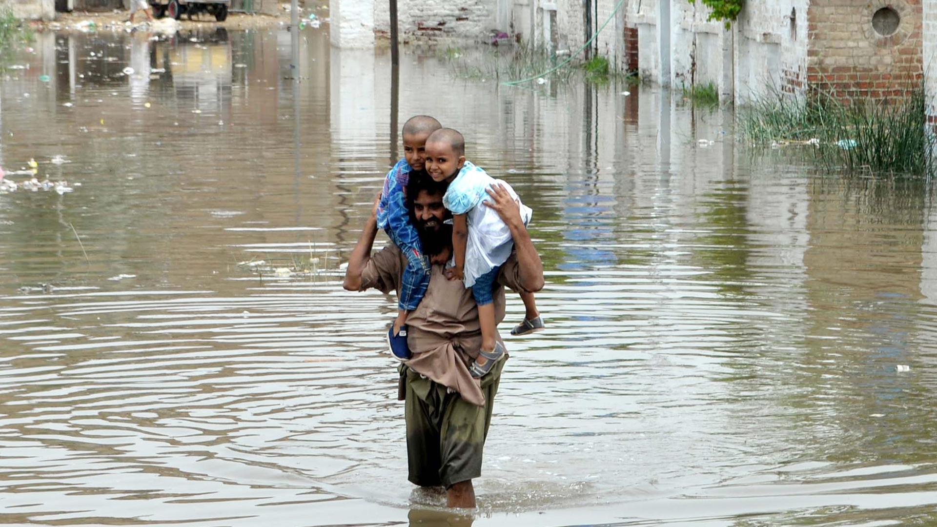 Der Mann im braunen Gewand steht auf einer völlig überfluteten Straße im Wasser. Er trägt seine zwei Kinder auf den Schultern. Im Hintergrund Häuserwände. Der Mann im braunen Gewand steht auf einer völlig überfluteten Straße im Wasser. Er trägt seine zwei Kinder auf den Schultern. Im Hintergrund Häuserwände.