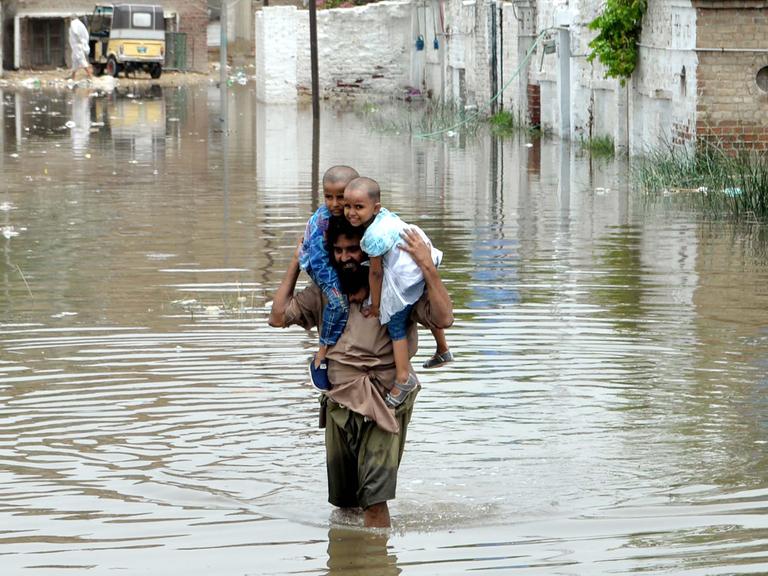 Der Mann im braunen Gewand steht auf einer völlig überfluteten Straße im Wasser. Seine zwei Kinder lachen. Im Hintergrund Häuserwände.