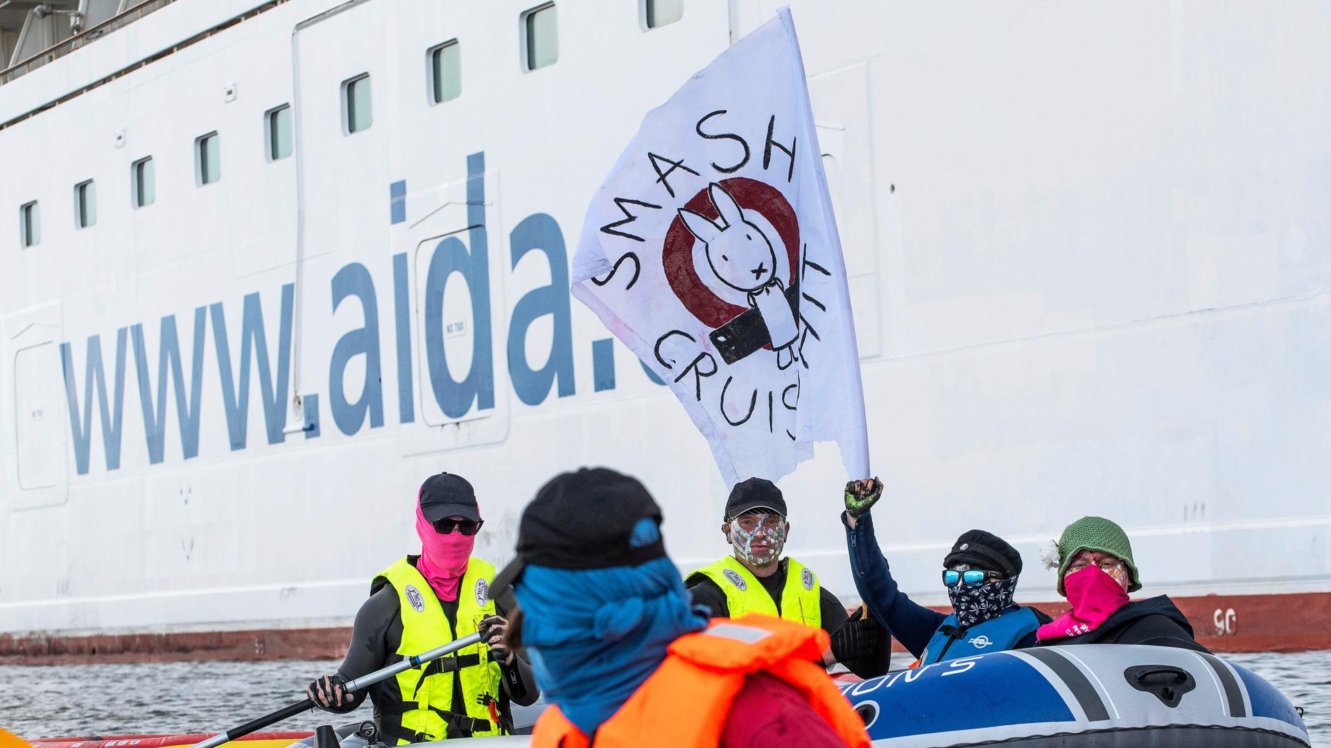 Mehrere Menschen mit Masken vor dem Gesicht sitzen in Schlauchbooten und paddeln um ein Kreuzfahrtschiff mit der Aufschrift "Aida" herum. Sie schwenken eine weiße Flagge mit der schwarzen Aufschrift "Smash Cruiseshit". So nennt sich die Aktivisten-Gruppe.