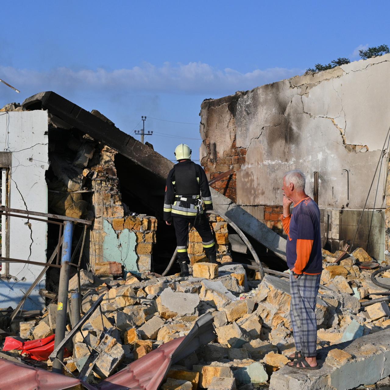 Ein Anwohner betrachtet die Zerstörungen nach einem russischen Raketenangriff. Er steht in den Trümmern eines Hauses. Im Hintergrund steht ein Feuerwehrmann. 