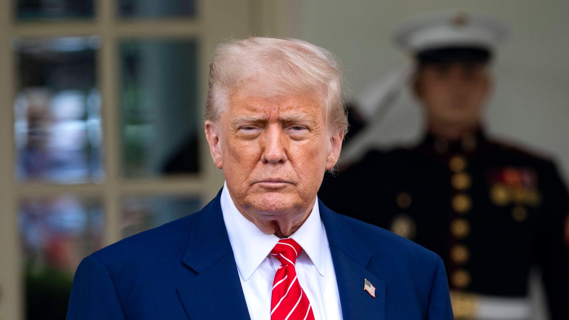 US-Präsident Donald Trump trägt einen blauen Anzug, im Hintergrund steht ein Soldat, der salutiert. Trump spricht mit Reportern vor dem Westflügel des Weißen Haus.