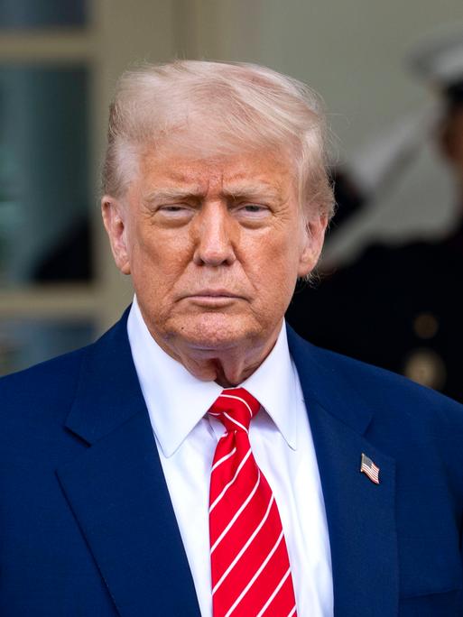 US-Präsident Donald Trump trägt einen blauen Anzug, im Hintergrund steht ein Soldat, der salutiert. Trump spricht mit Reportern vor dem Westflügel des Weißen Haus.