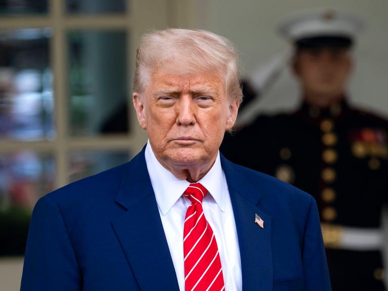 US-Präsident Donald Trump trägt einen blauen Anzug, im Hintergrund steht ein Soldat, der salutiert. Trump spricht mit Reportern vor dem Westflügel des Weißen Haus.