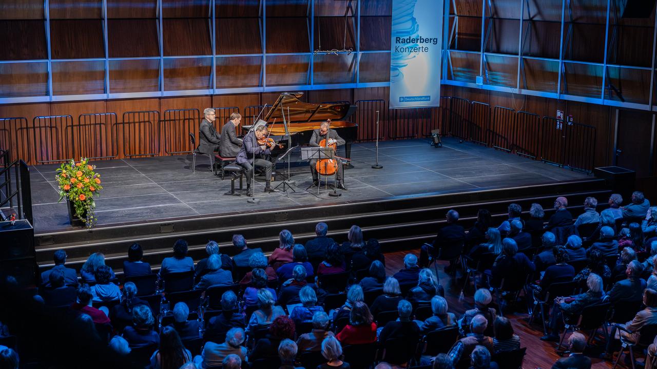 Blick auf ein männliches Klaviertrio, das auf einer Bühne konzertiert. An der Seite des Pianisten sitzt ein Notenwender. Der holzvertäfelte Saal ist blau erleuchtet.