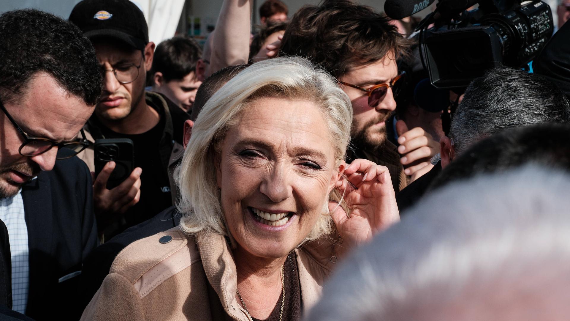 Die französische Rechtspopulistin Marine Le Pen läuft inmitten mehrerer Pressevertreter. Sie blickt zur Kamera und lacht.