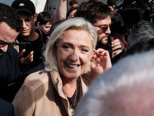 Die französische Rechtspopulistin Marine Le Pen läuft inmitten mehrerer Pressevertreter. Sie blickt zur Kamera und lacht.