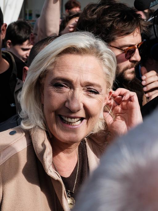 Die französische Rechtspopulistin Marine Le Pen läuft inmitten mehrerer Pressevertreter. Sie blickt zur Kamera und lacht.