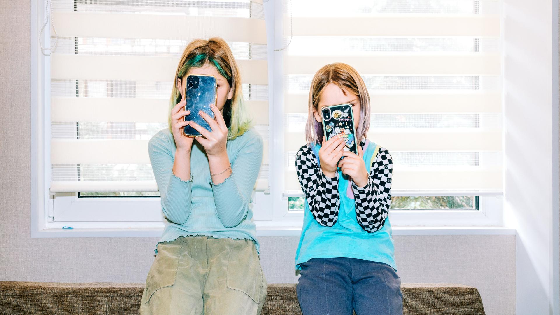 Zwei Kindernutzen jeweils ein Smartphone und verdecken damit ihre Gesichter. 