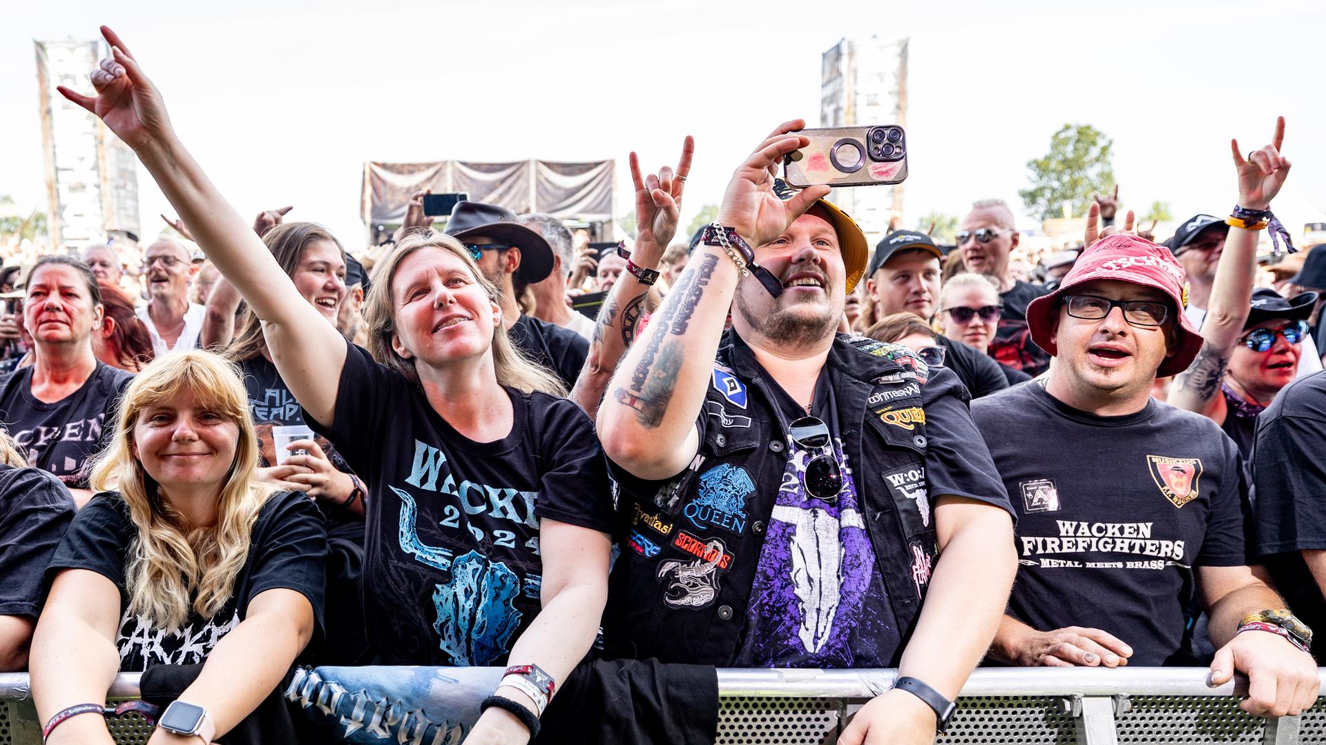 Lächelnde Frauen und Männer in schwarzen T-Shirts stehen vor einer Absperrung. Viele Shirts haben einen "Wacken"-Aufdruck. Eine Frau macht eine "Pommes-Gabel": sie streckt Zeigefinger und kleinen Finger hoch, die anderen Finger zur Handfläche gekrümmt. 