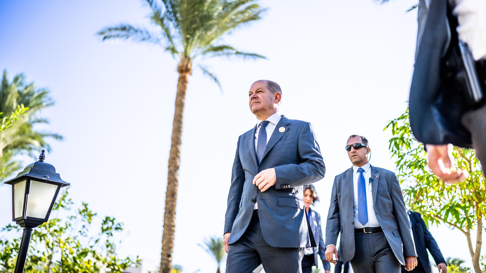 Olaf Scholz geht bei der UN-Weltklimakonferenz in Ägypten an einer Palme vorbei, hinter ihm folgt ein Personenschützer mit Sonnenbrille. Olaf Scholz geht bei der UN-Weltklimakonferenz in Ägypten an einer Palme vorbei, hinter ihm folgt ein Personenschützer mit Sonnenbrille.