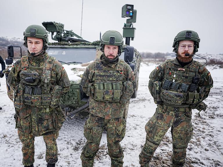 Drei Bundeswehr-Soldaten stehen im Schnee vor einem gepanzerten Fahrzeug.