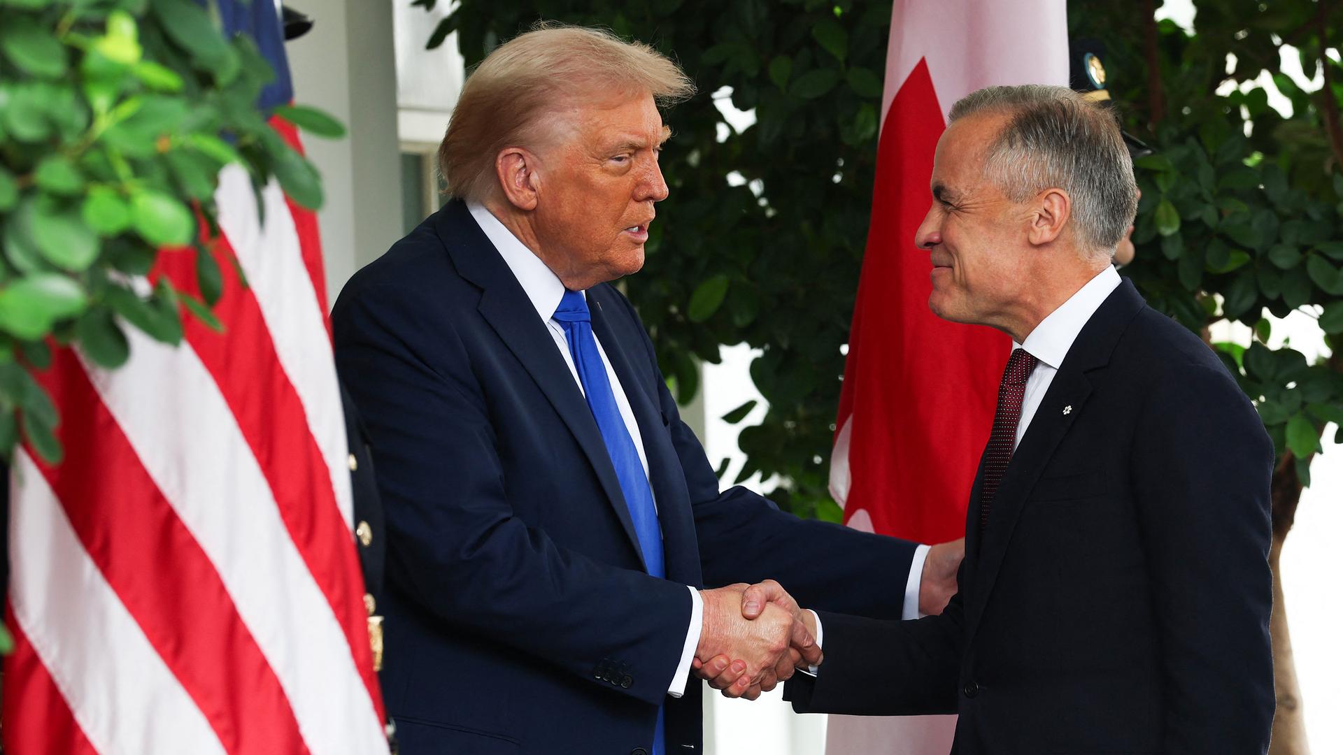 Donald Trump und Mark Carney schütteln einander die Hände. Sie stehen vor der amerikanischen und der kanadischen Flagge.